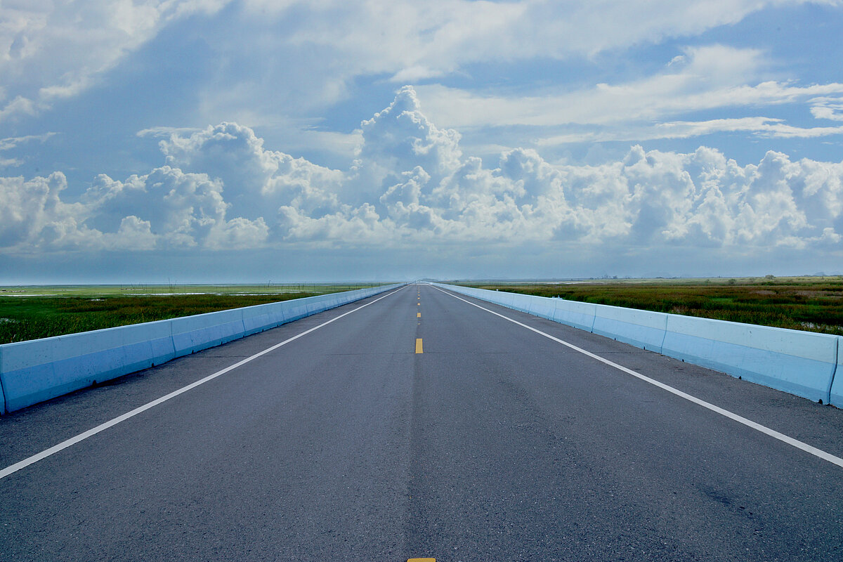 Leere gerade verlaufende Straße unter blau weißem Himmel.